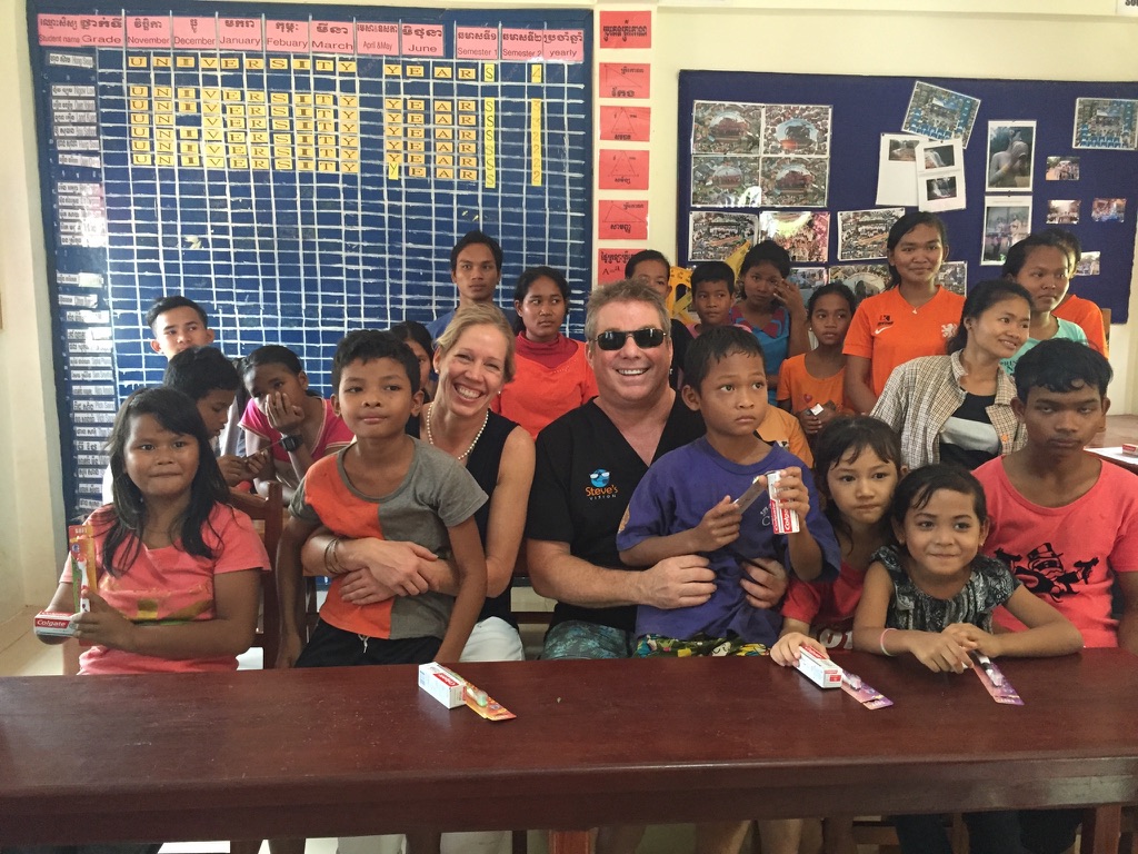 Steve Bender and an Oral Hygiene Initiative volunteer sit with a classroom of smiling students holding new toothbrushes and toothpaste