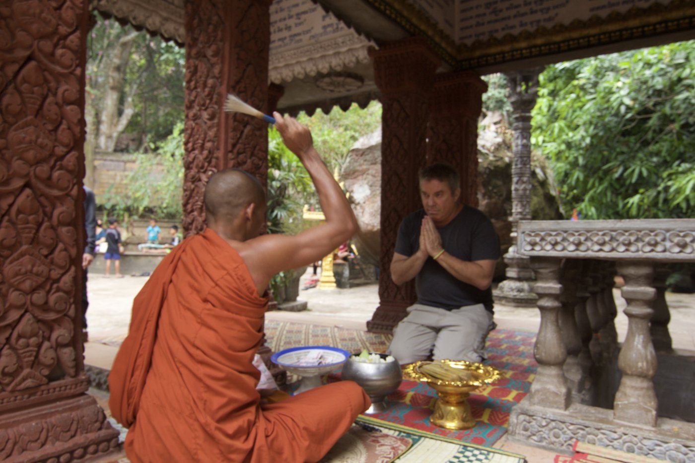 A monk in saffron robes sprinkles blessed water over Steve Bender, who kneels with hands pressed together in a carved temple pavilion