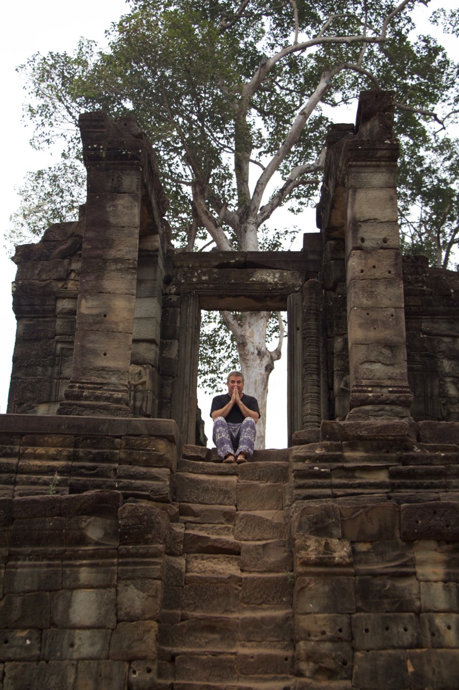 Steve Bender sits with hands pressed together in a temple doorway beneath a towering tree